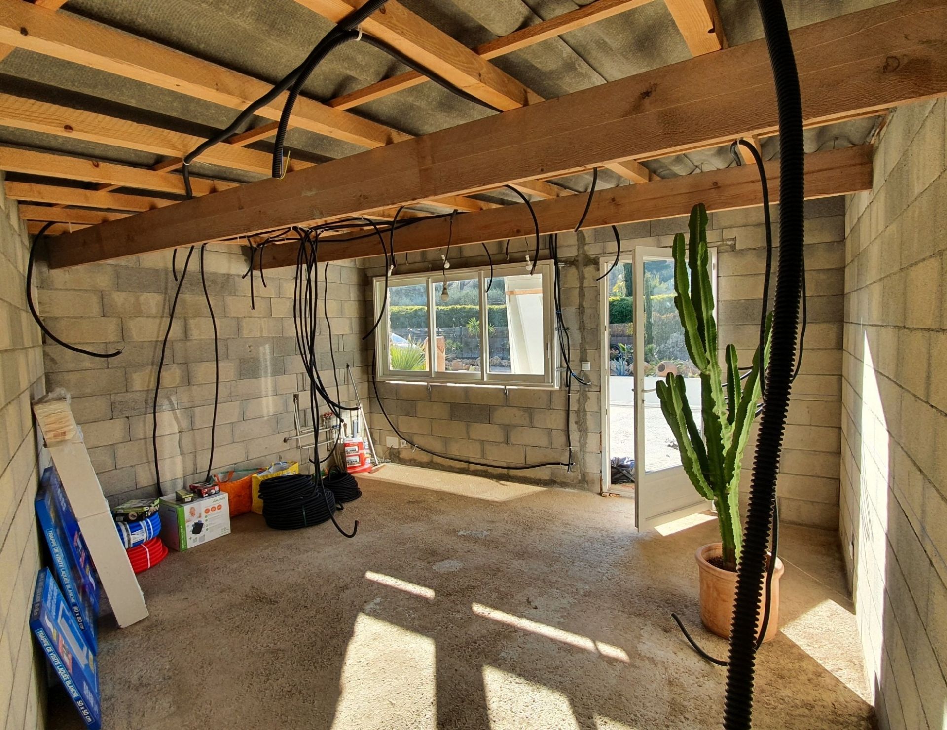 avant rénovation pièce Renov’Var Intérieur en construction avec murs en parpaings, poutre bois, installations électriques, fenêtres, cactus en pot et